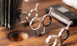 Close-up of shiny metal scissors handles in a wooden holder, with a round brush and an electric hair trimmer in the background at the Best Beauty Salon in Hisar, Haryana.