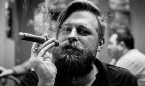 A man with a beard and mustache smokes a cigar, holding it near his face as smoke curls upwards. He wears a ring and a dark shirt. The blurred background hints at Haryana, with another person visible. The image is in black and white.