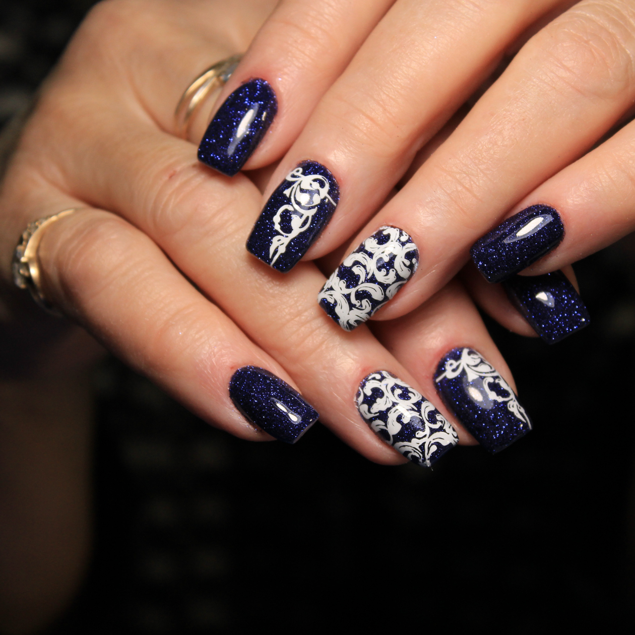 Hands with square-shaped nails painted dark blue and glittery, featuring intricate white floral designs on the middle and ring fingers. The person, possibly from Haryana, is wearing gold rings—nail art worthy of the Best Beauty Salon in Hisar.