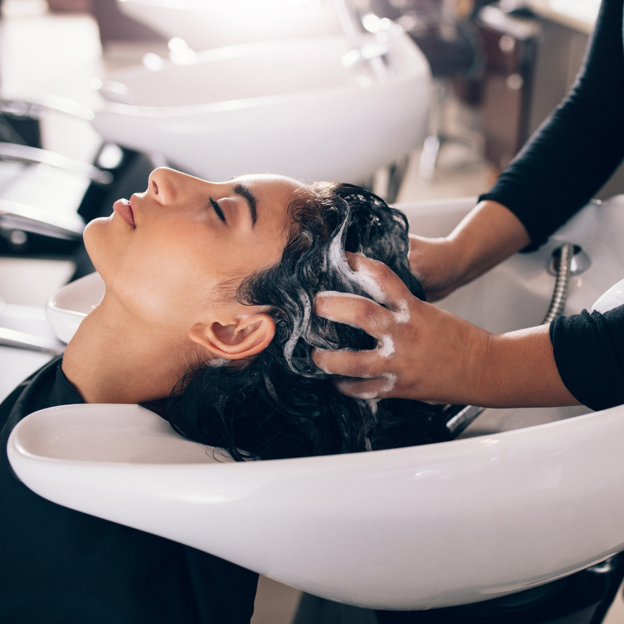 A person reclines at a salon sink while a hairstylist from the Best Beauty Salon in Hisar, Haryana washes their hair, massaging shampoo into their scalp as white suds form. The scene is bright and professional.