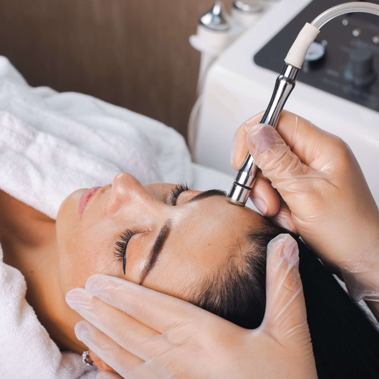 A woman lies with her eyes closed as a professional at the Best Beauty Salon in Hisar performs a facial treatment using a microdermabrasion device on her forehead in a serene spa setting.