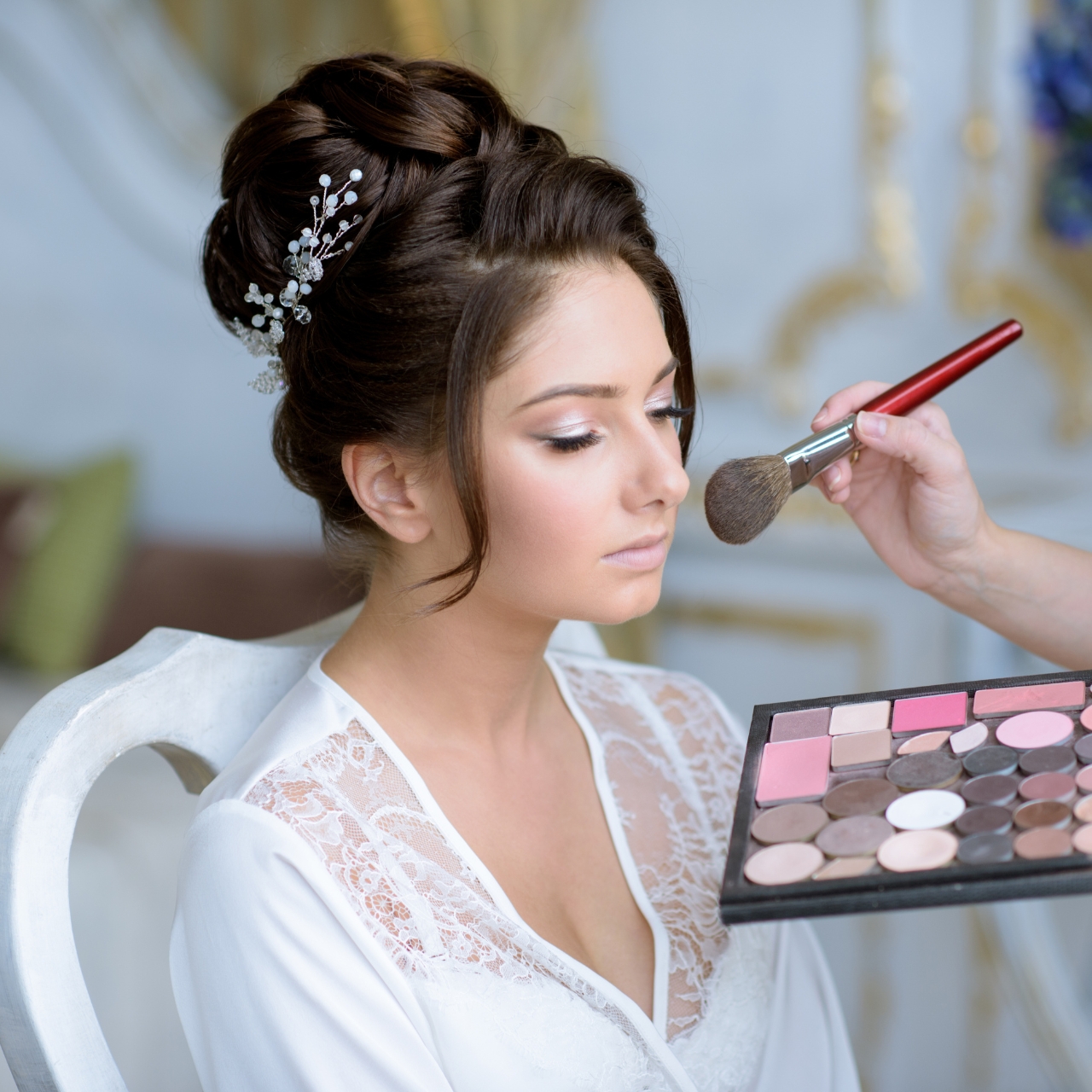 A woman with styled hair and a decorative hairpiece sits calmly as another person uses a brush and makeup palette—an experience youd find at the Best Beauty Salon in Hisar. She wears a white lace robe, looking serene and composed.